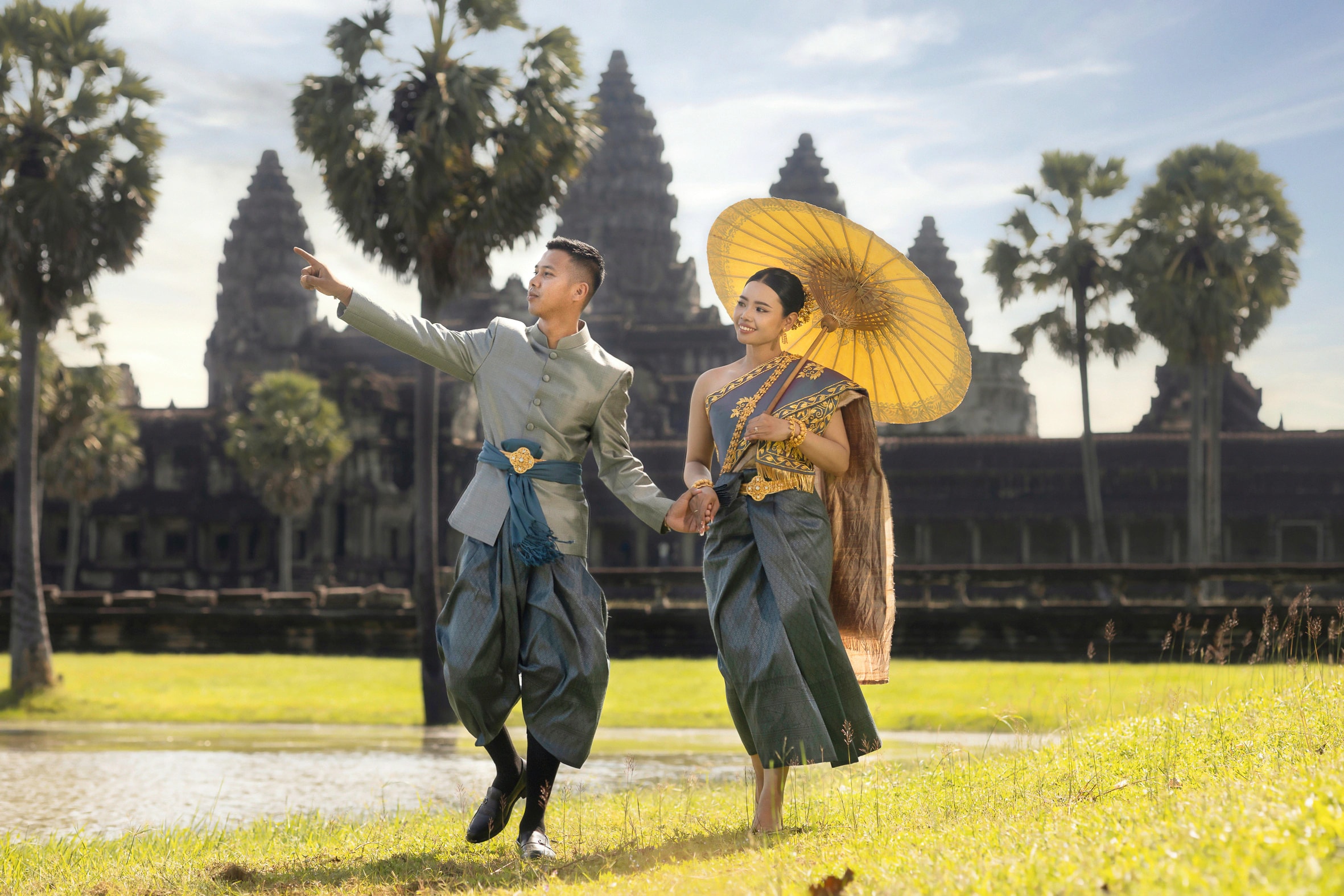 A couple in traditional Khmer dress walking hand in hand before Angkor Wat, the bride holding a gold parasol